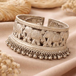 May include: Silver-toned cuff bracelet featuring a row of intricately carved elephants. The bracelet has a detailed design with small dangling bells along the bottom edge, creating a unique and eye-catching accessory.