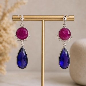 Op de afbeelding: Zilveren oorbellen met een ronde, magenta edelsteen en een diepblauwe, druppelvormige edelsteen. De oorbellen worden gepresenteerd op een gouden standaard.