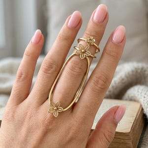 May include: A gold-toned metal ring featuring an elongated oval frame that spans across two fingers. The ring is adorned with three small, five-petalled flower motifs, each with intricate leaf-like cutouts, positioned at the top and bottom.