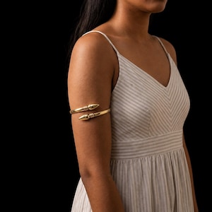 May include: A gold-toned metal arm cuff featuring a bypass design with two pointed, tapered ends. Each end is accented with three horizontal ridges near the tip. The arm cuff is worn on the upper arm, encircling it with a smooth, polished finish.
