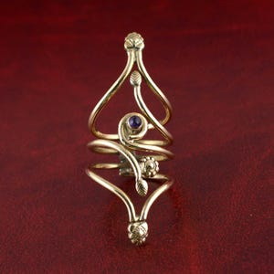 Può includere: Un anello dorato e ornato con un design a spirale. L'anello presenta una piccola gemma rotonda viola e accenti floreali e di foglie. L'anello ha una forma allungata unica e si avvolge attorno al dito.