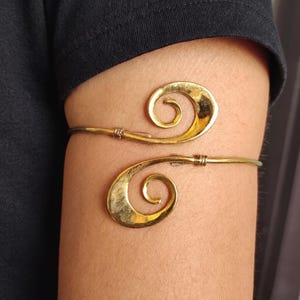 May include: Gold-coloured arm cuff with a double spiral design. The cuff is made of thin metal and wraps around the arm. The spirals have a textured finish, adding visual interest to the jewellery. The arm cuff is worn on the upper arm.