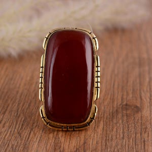 May include: A rectangular ring featuring a large, polished, deep red stone set in a gold-toned metal frame. The frame has a decorative, geometric design. The ring is displayed on a wooden surface, showcasing its rich color and design.