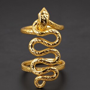 May include: A gold-toned ring featuring a snake design that wraps vertically across the finger. The snake has a textured, scaled surface and a defined head at the top, connecting two horizontal bands that form the base of the ring structure.
