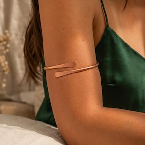 May include: A metallic copper-toned arm cuff featuring a simple, open-ended design with slightly flattened, tapered ends. The band wraps around the upper arm, showcasing a smooth, polished surface that reflects light across its curved structure.