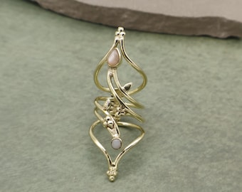 May include: A gold-colored, multi-finger ring with an ornate design. The ring features a series of swirling bands, small floral accents, and two oval-shaped, light-colored stones. The ring is set against a muted green background.