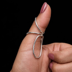 May include: A silver-colored metal splint ring designed to wrap around the thumb joint. The piece features a smooth, continuous wire construction that forms a circular band around the base of the thumb and an elongated loop extending downward.