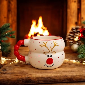 Könnte beinhalten: Eine weiße Keramik-Weihnachtstasse mit Rentiergesicht-Design, roten Punkten und rotem Griff. Die Tasse steht auf einer Holzoberfläche mit Kamin und Weihnachtsdekorationen im Hintergrund.