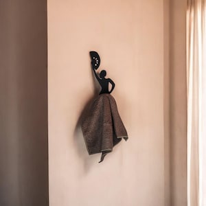 Può includere: Un gancio da parete in metallo nero a forma di ballerina di flamenco con un ventaglio. Un asciugamano marrone a fantasia è drappeggiato sulla gonna della ballerina. Il gancio è montato su una parete color pesca chiaro, con una tenda a destra.