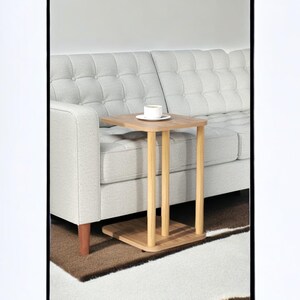 Könnte beinhalten: Kleiner Beistelltisch aus hellem Holz mit quadratischer Tischplatte und drei zylindrischen Beinen. Der Tisch steht neben einem hellgrauen Sofa. Eine weiße Tasse mit Untertasse steht auf dem Tisch.