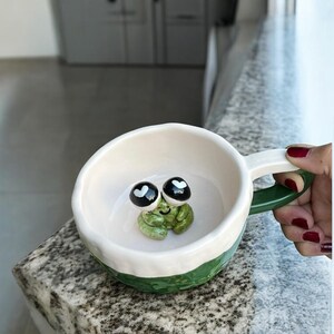 Könnte beinhalten: Eine weiße Keramiktasse mit grünem Boden und Griff, mit einer skurrilen Froschfigur im Inneren. Der Frosch hat große schwarze Augen mit weißen herzförmigen Akzenten. Die Tasse wird von einer Hand mit rotem Nagellack gehalten.