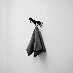 Puede incluir: Un gancho de pared de metal negro con forma de perro, montado en una pared blanca. Una toalla gris doblada cuelga del gancho. El diseño del gancho es minimalista y moderno, adecuado para un baño o una entrada.