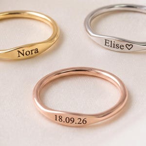 Könnte beinhalten: Drei dünne Metallringe mit flachen, gravierten Oberflächen. Ein goldener Ring ist mit dem Namen Nora graviert, ein silberner Ring mit dem Namen Elise und einem Herzsymbol, und ein roségoldener Ring trägt die Gravur des Datums 18.09.26.
