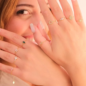 Puede incluir: Una colección de anillos de oro con varios diseños, incluyendo corazones, flores y piedras preciosas. Los anillos se muestran en dedos con uñas largas y cuidadas. La joyería es delicada y elegante, adecuada para el uso diario o ocasiones especiales.