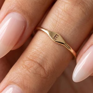 Peut inclure: Une fine bague en or dotée d'une petite plaque plate de forme ovale sur le devant. Le centre de la plaque est gravé de la lettre majuscule E. L'anneau de la bague est lisse, poli et circulaire, conservant une épaisseur constante partout.