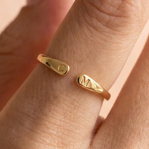 May include: A gold-toned open ring featuring two tapered ends, each engraved with a single letter. One end displays the letter C, while the other end displays the letter M. The band has a smooth, polished finish and a simple, adjustable design.