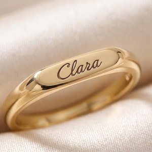 May include: A polished gold-toned ring featuring a flat, elongated rectangular top surface. The name Clara is engraved in a cursive script font across the center of the flat surface. The band has a smooth, rounded finish with a reflective metallic sheen.