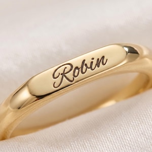 May include: A polished gold-toned ring featuring a smooth, flat surface on the top band. The name Robin is engraved in a dark, cursive script font across the center of the flat section, creating a clear contrast against the metallic gold finish.