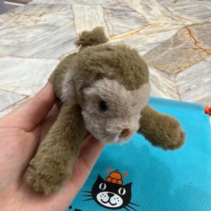 May include: A plush, brown and tan stuffed animal seal with a small tail and outstretched paws. The seal is held in front of a blue bag with the text "JELLYCAT LONDON" and a black cat logo.