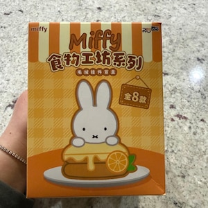 May include: A box featuring the character Miffy, with text in English and Chinese. The box has a cartoon image of Miffy on a dessert. The box is orange and yellow with a checkered pattern.