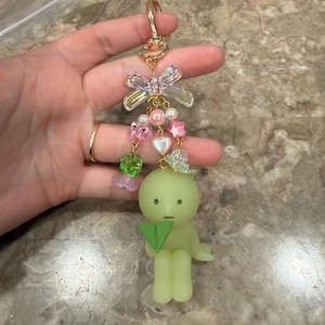 May include: A whimsical keychain featuring a light green figurine holding a green paper airplane. The keychain has a gold-colored clasp and is adorned with pink, white, and clear beads, and a clear bow.