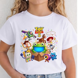 May include: A white t-shirt featuring a colorful graphic print of Toy Story 5 characters including Buzz Lightyear, Woody, Jessie, Bullseye, and other animated figures surrounding a green frog in a cardboard box with confetti accents.