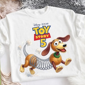 May include: A white t-shirt featuring a graphic of the Slinky Dog character from Toy Story. Above the character, the text reads Disney Pixar Toy Story 5. The dog has a brown body, a metal spring midsection, and a green collar with a gold tag.