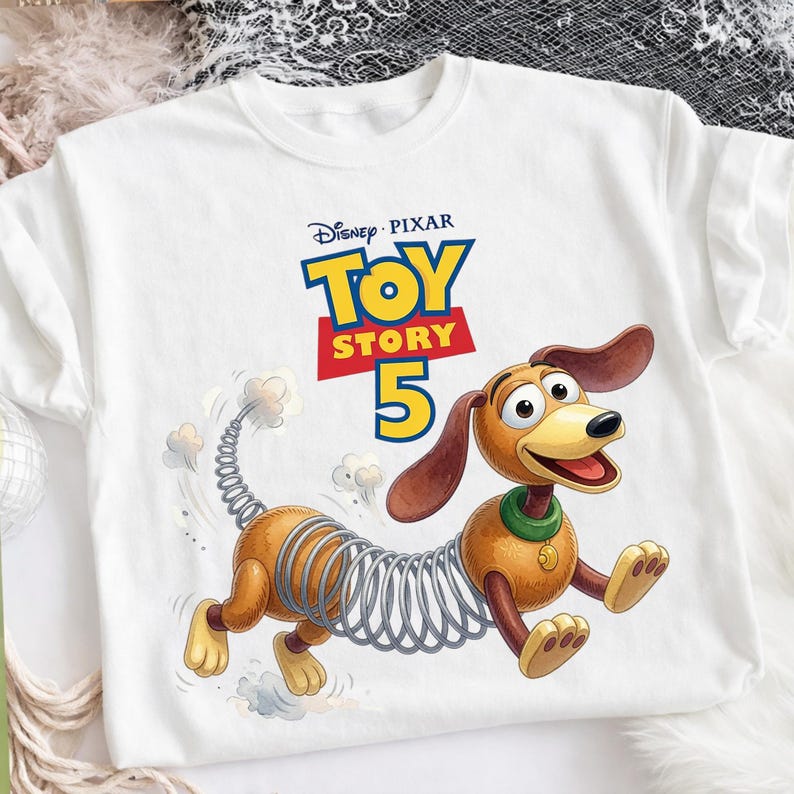 May include: A white t-shirt featuring a graphic of the Slinky Dog character from Toy Story. Above the character, the text reads Disney Pixar Toy Story 5. The dog has a brown body, a metal spring midsection, and a green collar with a gold tag.