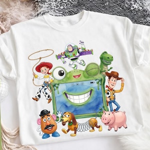 May include: A white t-shirt featuring a colorful graphic of Toy Story characters including Woody, Buzz Lightyear, Jessie, Rex, Slinky Dog, Mr. Potato Head, and Hamm surrounding a central green frog-shaped tablet with a wide smiling mouth.