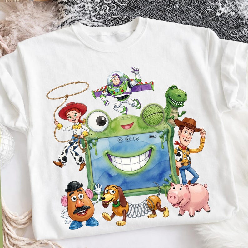 May include: A white t-shirt featuring a colorful graphic of Toy Story characters including Woody, Buzz Lightyear, Jessie, Rex, Slinky Dog, Mr. Potato Head, and Hamm surrounding a central green frog-shaped tablet with a wide smiling mouth.