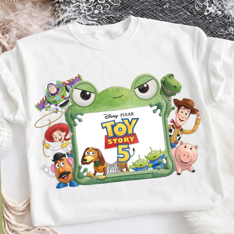 May include: A white short-sleeved t-shirt featuring a graphic print of Disney Pixar Toy Story characters including Buzz Lightyear, Woody, Jessie, Mr. Potato Head, Slinky Dog, Rex, Hamm, and Aliens surrounding a green frog-shaped frame with text.