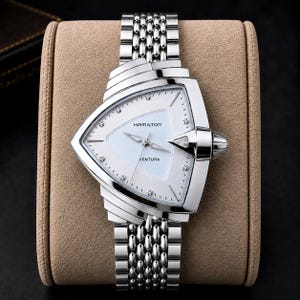 May include: A silver-tone Hamilton Ventura watch with a distinctive triangular case and a white dial. The watch features a metallic link bracelet, silver hands, and small dot hour markers. The brand name Hamilton and the model name Ventura are printed on the dial.