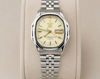 Puede incluir: Un reloj automático Seiko 5 Superior de acero inoxidable plateado con esfera color crema texturizada, marcadores de hora y manecillas doradas. El reloj cuenta con una ventana de día y fecha en la posición de las tres, brazalete de eslabones metálicos y resistencia al agua de 5 bares.