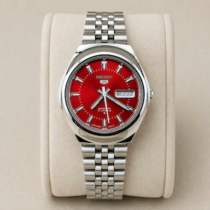 Op de afbeelding: Een roestvrijstalen Seiko 5 automatische polshorloge met een felrode wijzerplaat. Het horloge heeft een dag- en datumweergave, zilveren wijzers en een bijpassende roestvrijstalen armband. Het horloge wordt getoond op een crèmekleurig kussen.