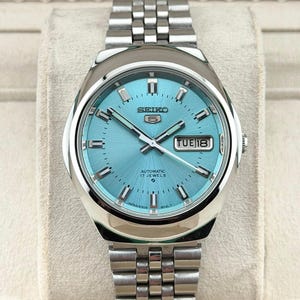Puede incluir: Un reloj de pulsera automático Seiko 5 plateado con esfera azul claro. El reloj presenta una visualización de día y fecha, agujas plateadas y una correa de acero inoxidable. La esfera del reloj tiene el texto "SEIKO 5 AUTOMATIC 17 JEWELS".
