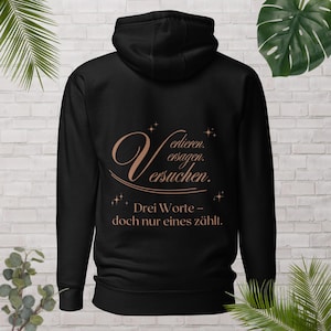 Könnte beinhalten: Schwarzer Kapuzenpullover mit kupferfarbenem deutschen Text: "Verlieren. versagen. versuchen. Drei Worte - doch nur eines zählt". Das Design beinhaltet dekorative Sterne. Ein lässiges Kleidungsstück.