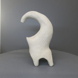 May include: A white ceramic sculpture with an abstract design, featuring a curved, horn-like shape. The textured sculpture stands on two legs. The artwork is set against a grey background, highlighting its form and design.