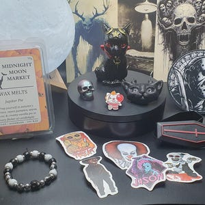 May include: A selection of Halloween-themed merchandise, including a black cat ornament, a skull, stickers, a bracelet, and wax melts. The wax melts are labelled "Midnight Moon Market" and "Jupiter Pie". The stickers feature horror movie characters.