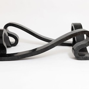 May include: A black granite sculpture with a flowing, ribbon-like design. The sculpture features two curved, intersecting elements, creating an abstract form. The polished surface reflects light, highlighting the dark colour and smooth texture.