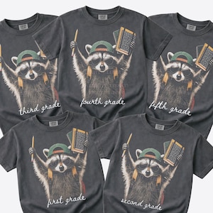 May include: Five charcoal grey t-shirts featuring a graphic of a raccoon wearing sunglasses and a green baseball cap, holding a pencil and a book. Each shirt displays a different grade level text: first grade, second grade, third grade, fourth grade, fifth grade.