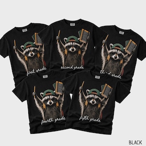 May include: Five black t-shirts featuring a graphic of a raccoon wearing sunglasses and a green baseball cap, holding a pencil and a notebook. Each shirt displays the text first grade, second grade, third grade, fourth grade, or fifth grade.