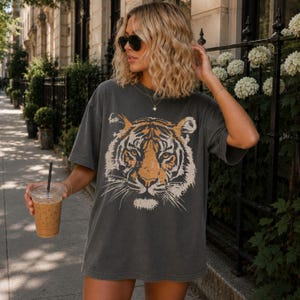 May include: A charcoal grey short-sleeve t-shirt featuring a large, detailed graphic print of a tiger face in shades of orange, black, and white. The shirt has a crew neckline and a relaxed, loose fit, showcasing a faded, worn-in appearance.