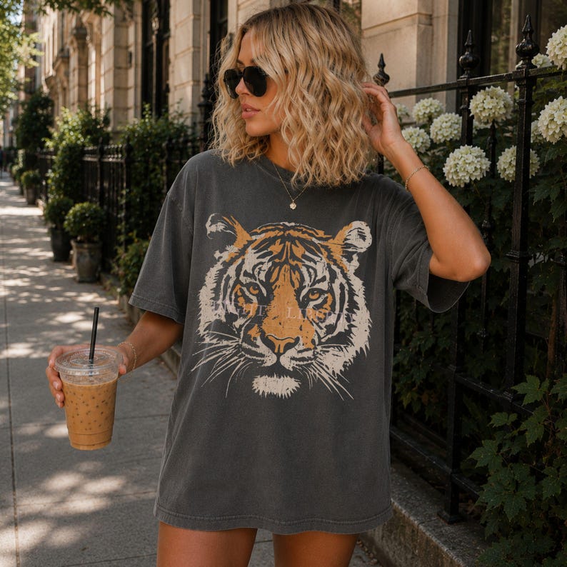 May include: A charcoal grey short-sleeve t-shirt featuring a large, detailed graphic print of a tiger face in shades of orange, black, and white. The shirt has a crew neckline and a relaxed, loose fit, showcasing a faded, worn-in appearance.