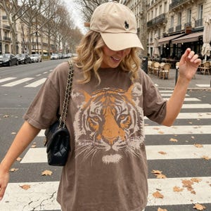 May include: A beige baseball cap with a small black embroidered logo and a brown short-sleeved t-shirt featuring a large orange and cream tiger face graphic print on the front, paired with a black quilted shoulder bag with a gold chain strap.