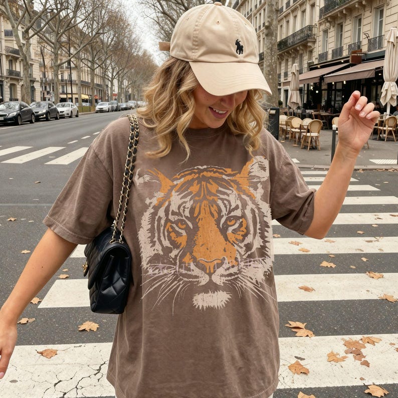 May include: A beige baseball cap with a small black embroidered logo and a brown short-sleeved t-shirt featuring a large orange and cream tiger face graphic print on the front, paired with a black quilted shoulder bag with a gold chain strap.