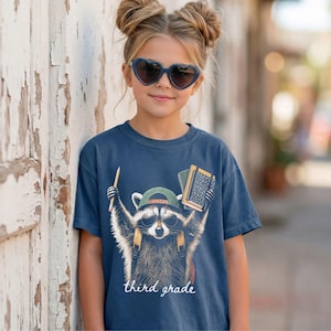May include: A navy blue t-shirt featuring a graphic of a raccoon wearing sunglasses and a green baseball cap. The raccoon holds a yellow pencil and a notebook. Below the graphic, the text reads third grade in a white cursive script font.