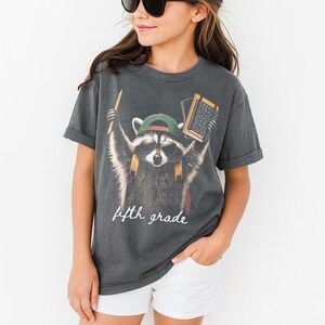 May include: A charcoal grey short-sleeved t-shirt featuring a graphic of a raccoon wearing sunglasses and a green baseball cap. The raccoon holds a yellow pencil and a notebook. The text "fifth grade" is printed in white script below the raccoon.
