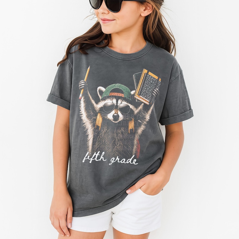 May include: A charcoal grey short-sleeved t-shirt featuring a graphic of a raccoon wearing sunglasses and a green baseball cap. The raccoon holds a yellow pencil and a notebook. The text "fifth grade" is printed in white script below the raccoon.