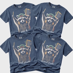 May include: Four blue short-sleeve t-shirts featuring a graphic of a raccoon wearing sunglasses and a green cap, holding a pencil and books. The shirts are labelled with the text pre-k, kindergarten, first grade, and second grade in white script.