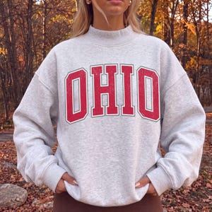 May include: A light gray heathered crewneck sweatshirt featuring the word OHIO printed in large, distressed red block letters with a white outline. The garment has long sleeves, a ribbed mock neck collar, and ribbed cuffs at the wrists and hem.